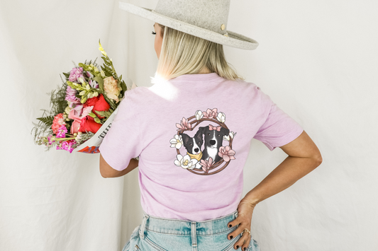 women with back turned, her shirt is lilac with a portrait of two black and white dogs one it. surrounding the dogs is a branch wreath with pink and white magnolias.
